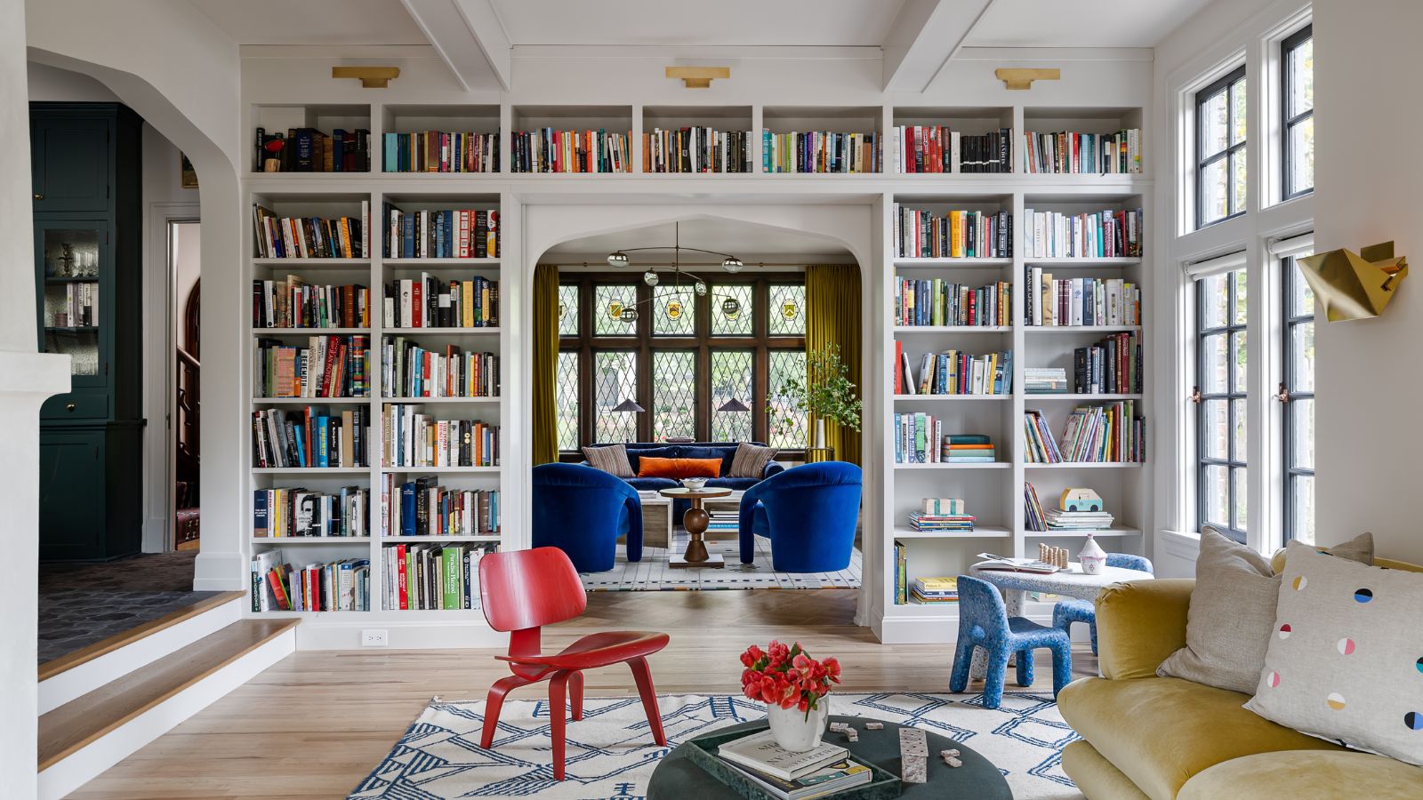 Living room with white painted walls, floor-to-ceiling bookcase with books, yellow velvet sofa, red chair on a rug, looking into another living room with blue armchairs and a blue sofa