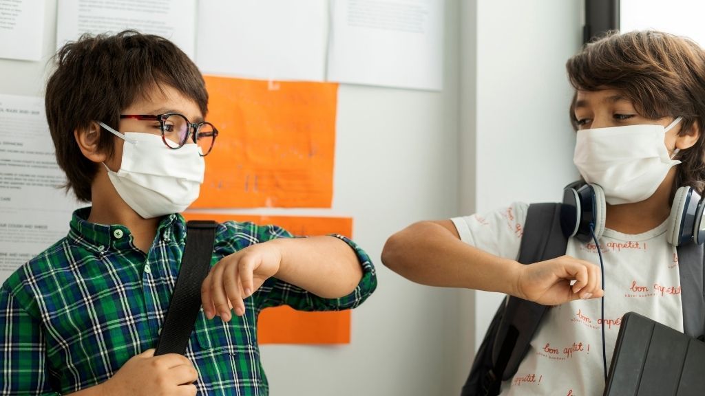 two boys in face masks bump elbows while standing near a wall in school