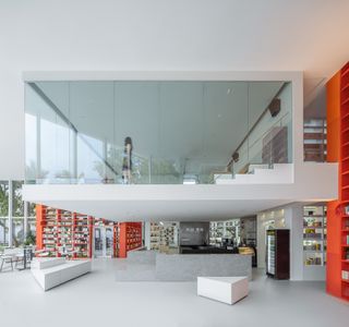 Interior room, white floor, walls and ceiling, orange shelving units, suspended glass viewing gallery in the centre, windows to the left with a surrounding area view of trees, counter top, white triangular seating, vending machine