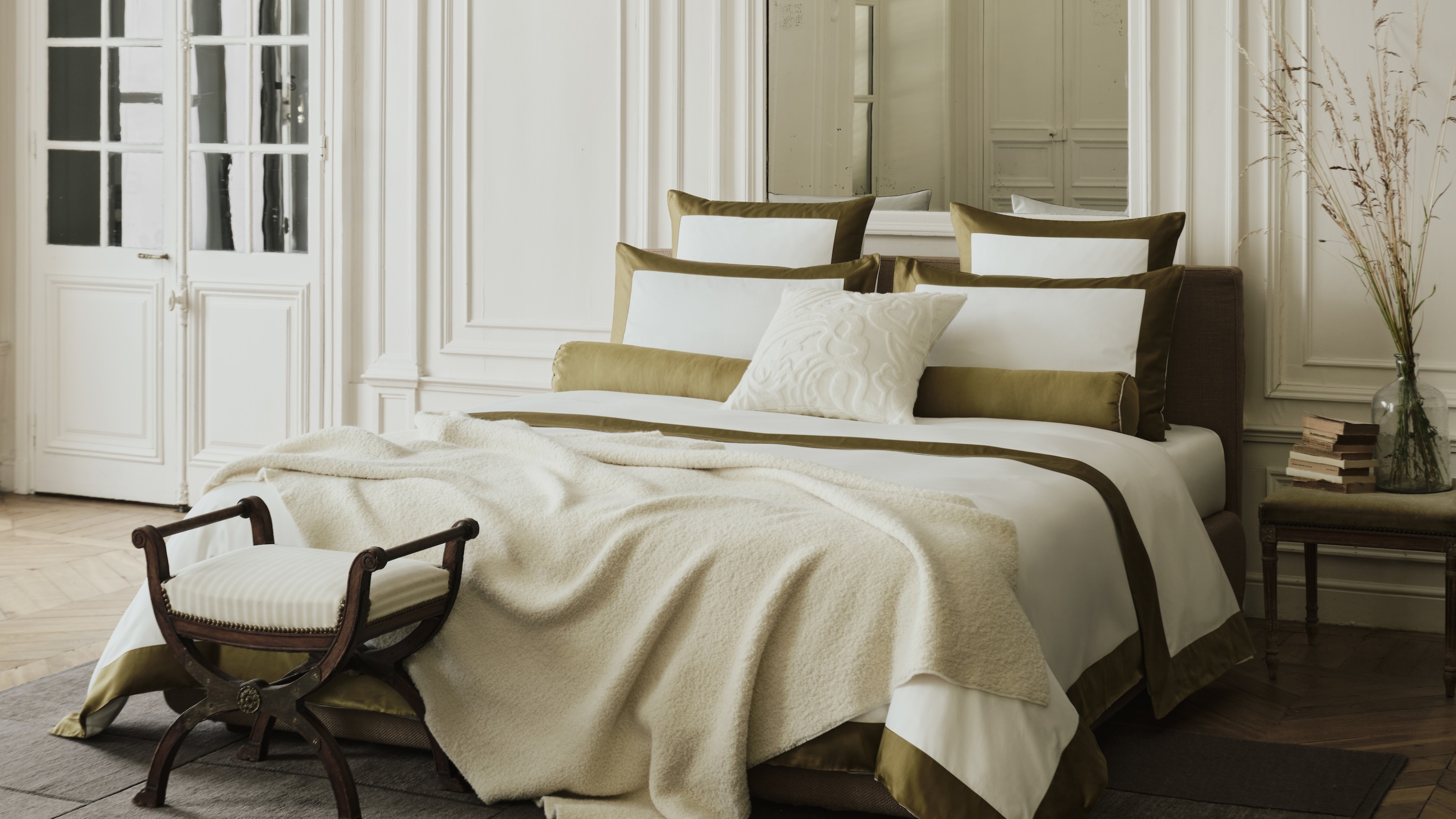 Image of a large, luxurious-looking bed in an open bedroom. The bedding is white with a dark green trim, and there are seven decorative pillows on the bed.