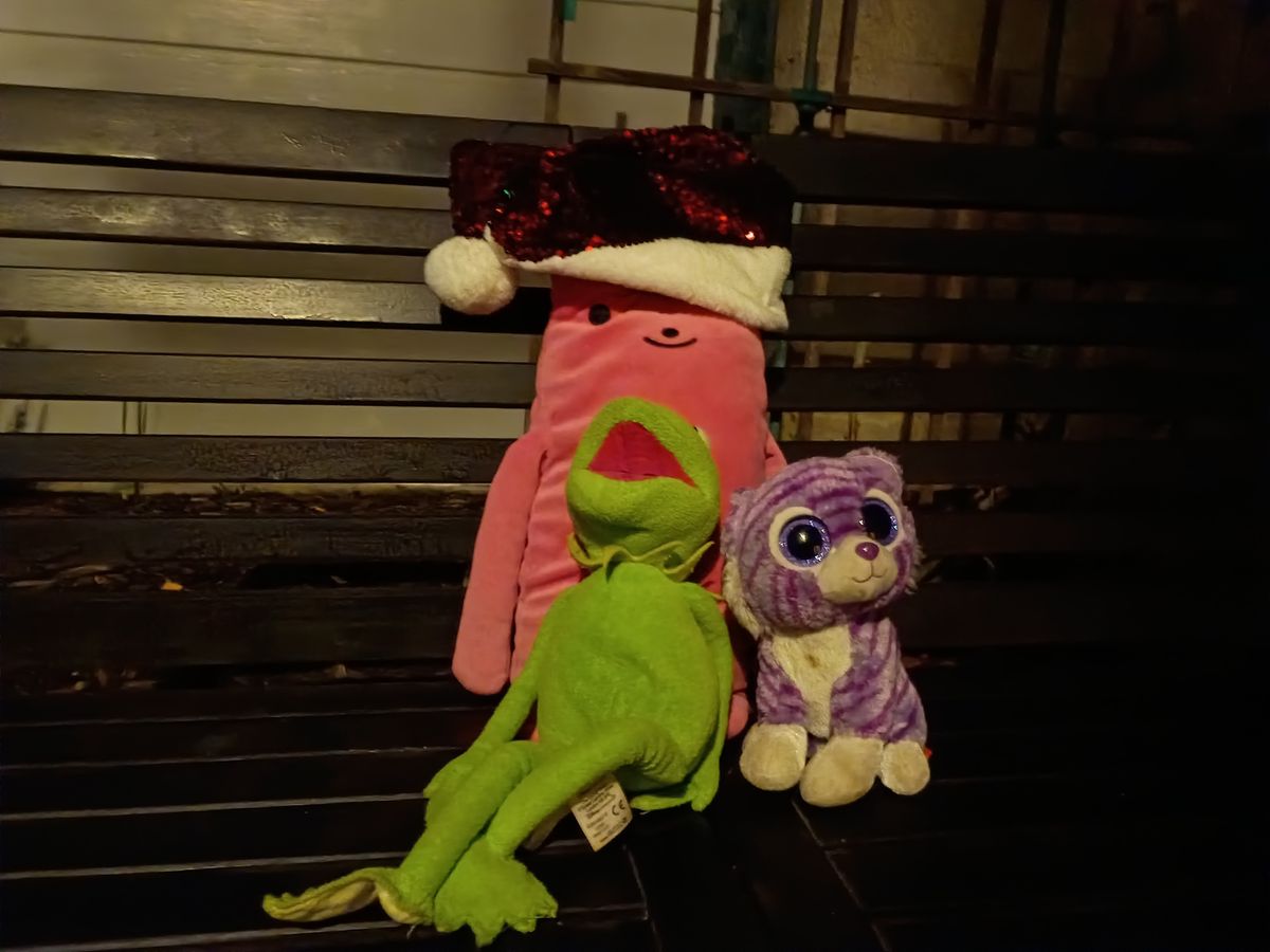 three children's soft toys placed on a wooden public bench outside late at night in artificial street light