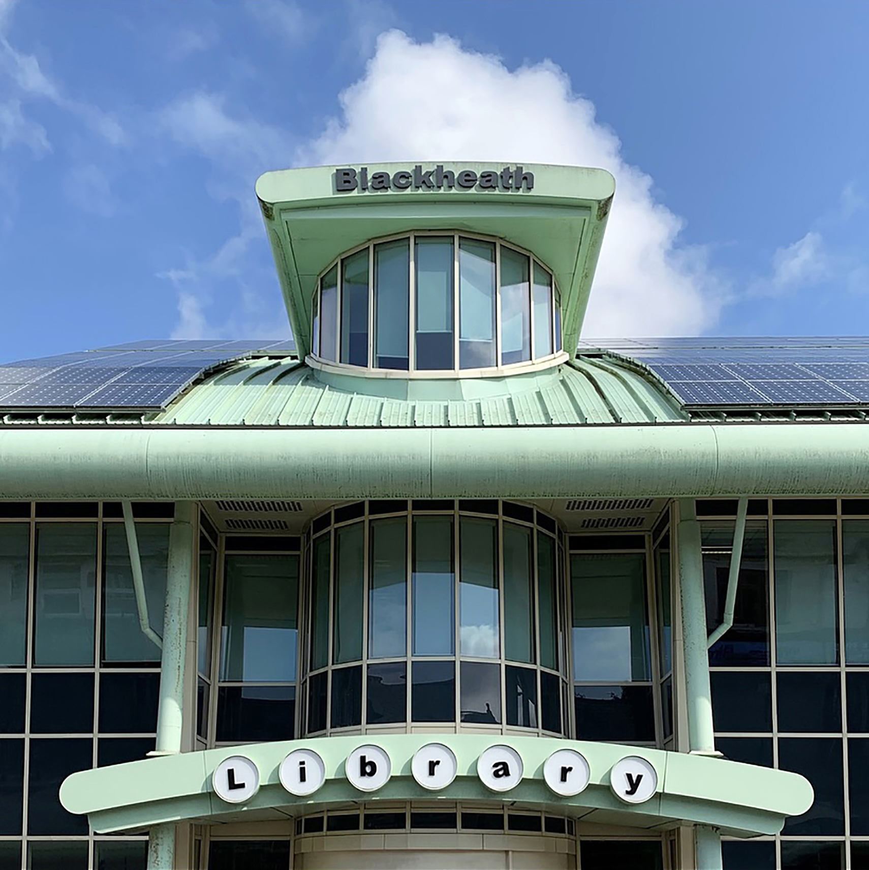 A pale green art deco building topped with a curved window and individually mounted letters spelling "Library" against a partly cloudy sky.