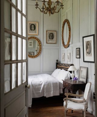 A doorway view into a French style bedroom with eclectic wall art on panelled walls, a black bed, wooden beside table and wooden upholstered chair