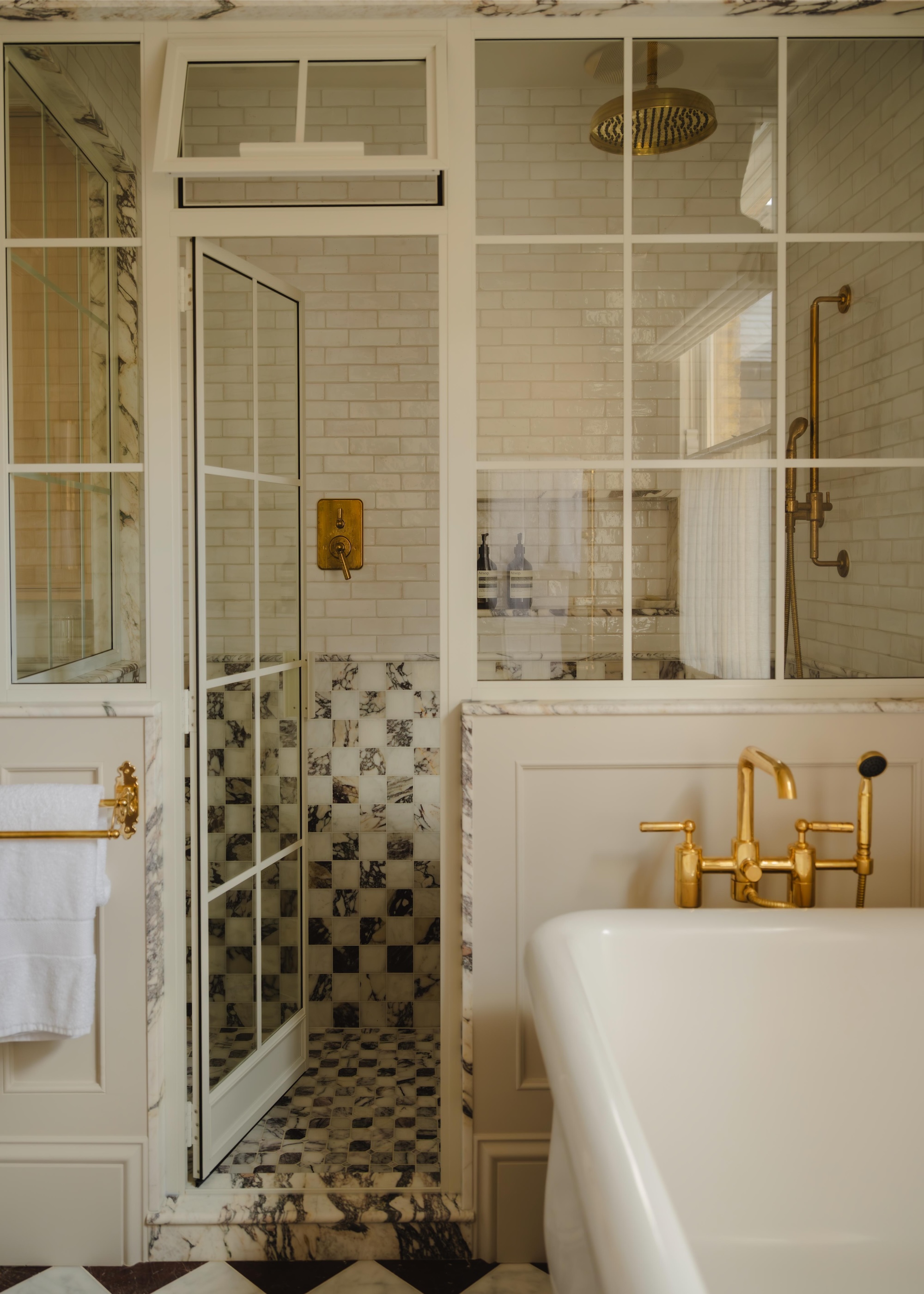A classic bathroom with neutral brick shaped tiles on the top half of the wall and a checquerboard style flooring, shown with a roll top bath