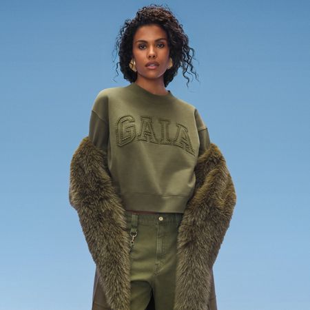 a model wears a gap sweatshirt with a faux fur coat and cargo pants in front of a plain backdrop