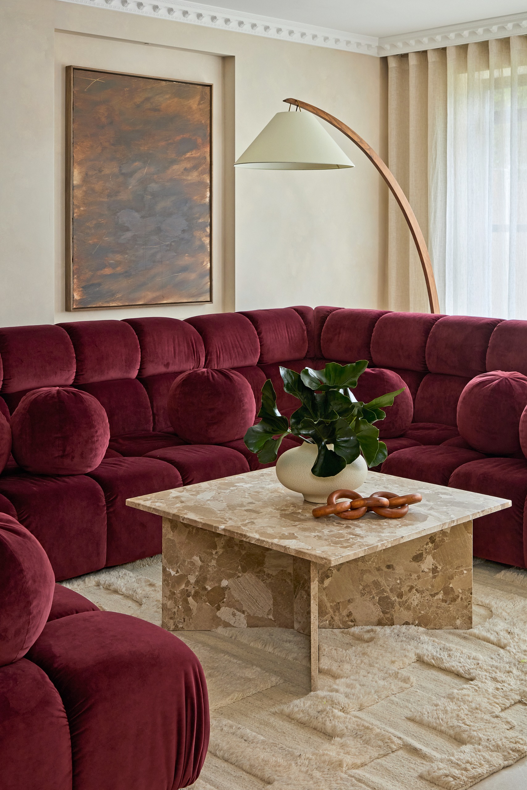 Cream living room with burgundy U-shaped sofa, beige marble coffee table, beige textured rug, curved wood floor lamp and bronze abstract artwork