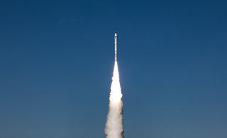 rocket blasts off atop a white plume with blue sky behind