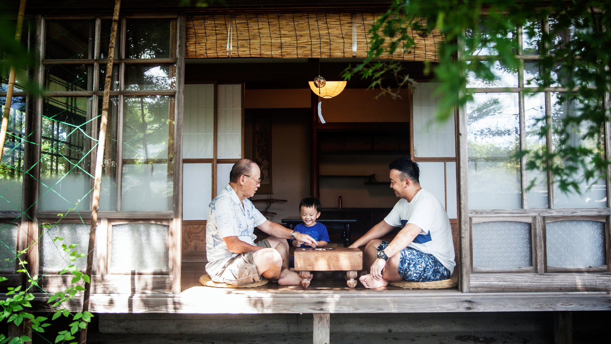 Japanese males and child sat on floor outside home with bamboo blind and planted screens