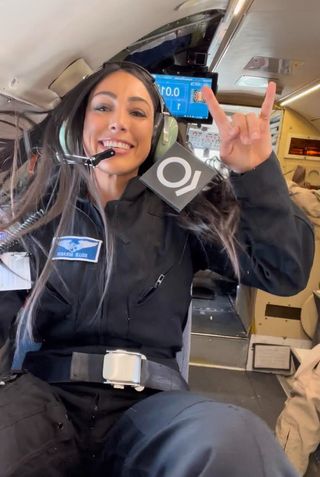 a woman wearing a flight suit weightless on an airplane gives the 'rock on' hand signal