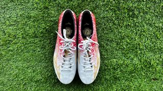 Red and white football boots with white laces and a gold lightning flash on the outside of the boot sit on top of green artificial grass. Mizuno Alpha Made in Japan