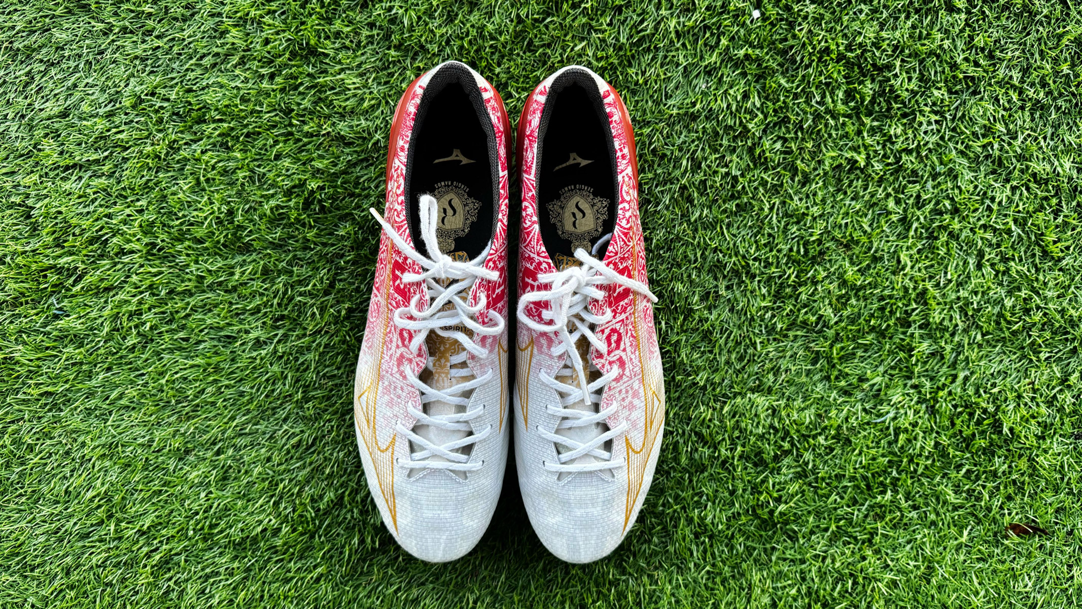 Red and white football boots with white laces and a gold lightning flash on the outside of the boot sit on top of green artificial grass. Mizuno Alpha Made in Japan