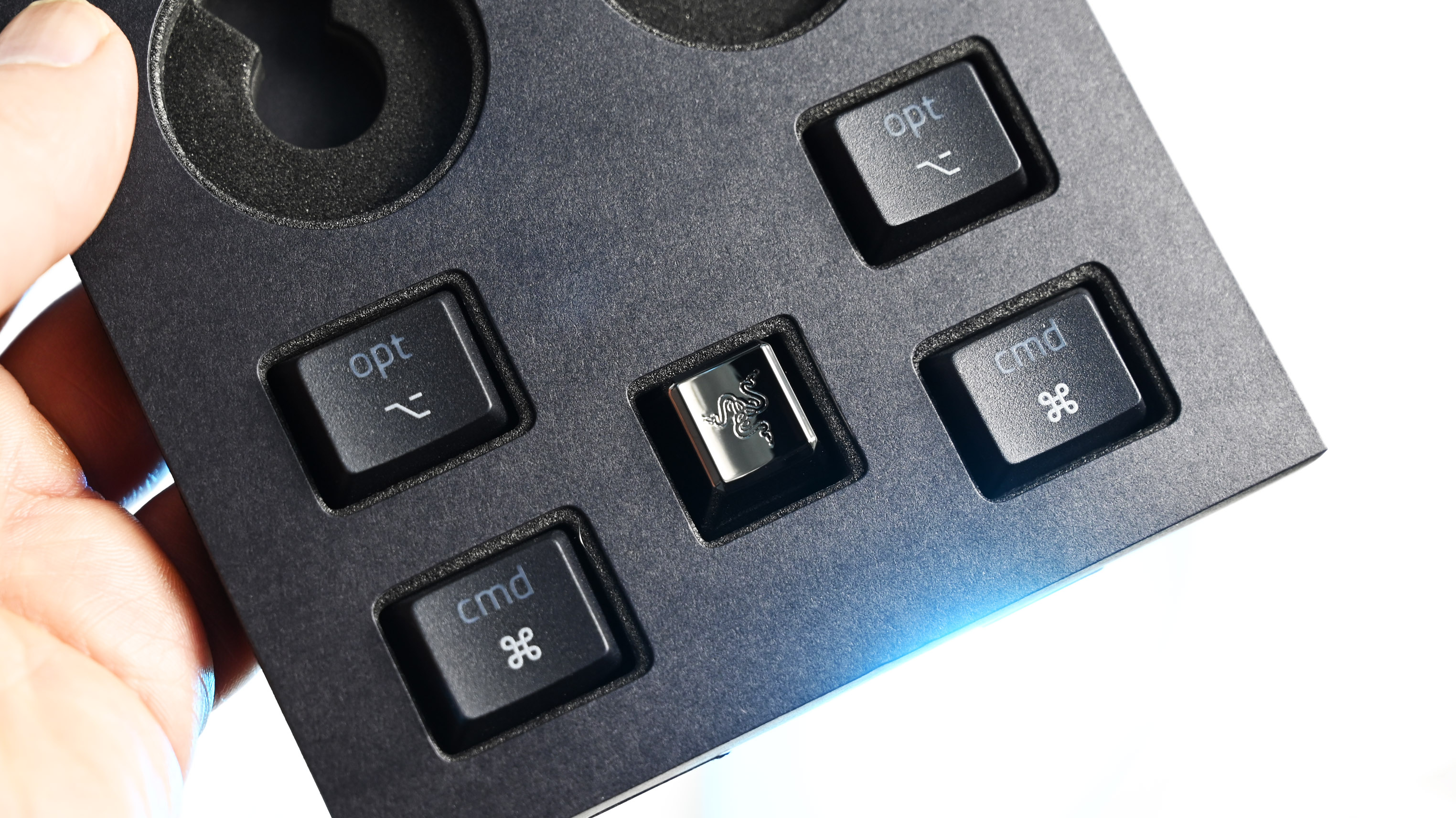 Close-up of a foam tray holding extra keycaps, including two Mac "cmd" keys, two "opt" keys, and a metallic chrome keycap with the Razer logo.
