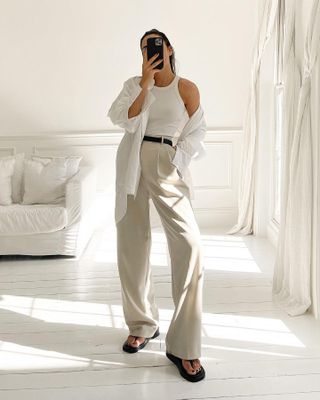 British fashion influencer Jessica Skye poses for a mirror selfie wearing a chic outfit formula with a white racerback tank top, open white button-down shirt, black belt, tan trousers, and black flip-flop sandals