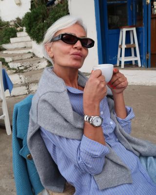 @greceghanem with natural skin, smiling at the camera, drinking a cup of coffee outdoors