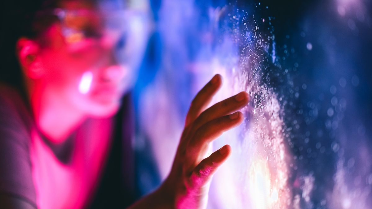 An out of focus worker putting their hand up to a glowing wall with data points on it, representing AI. Decorative: the worker is lit in punk light while the wall is purple, black, and bright orange.