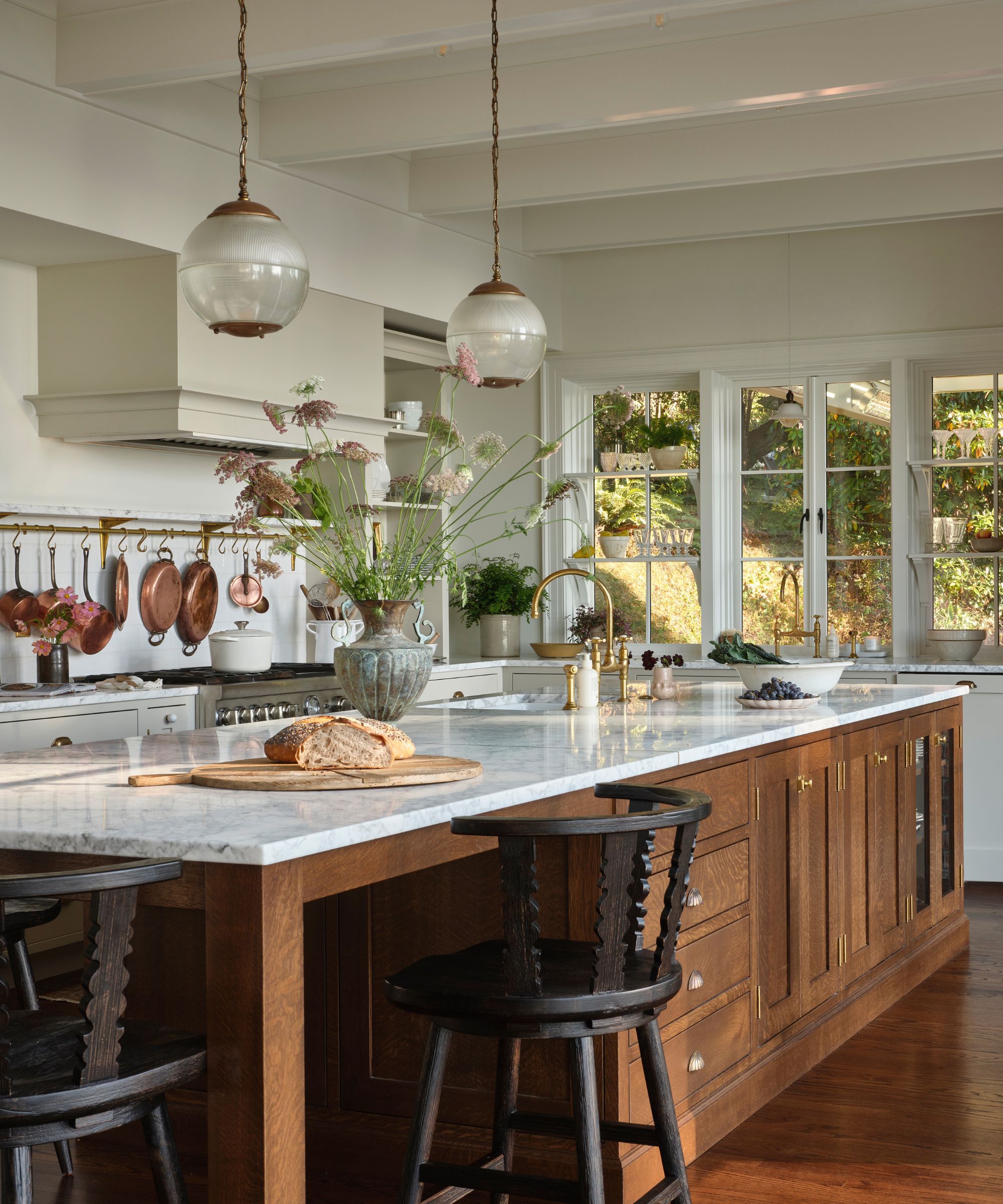 A spacious, sunlit kitchen featuring a large oak island with a marble countertop, black spindle stools, and vintage-style glass pendant lights