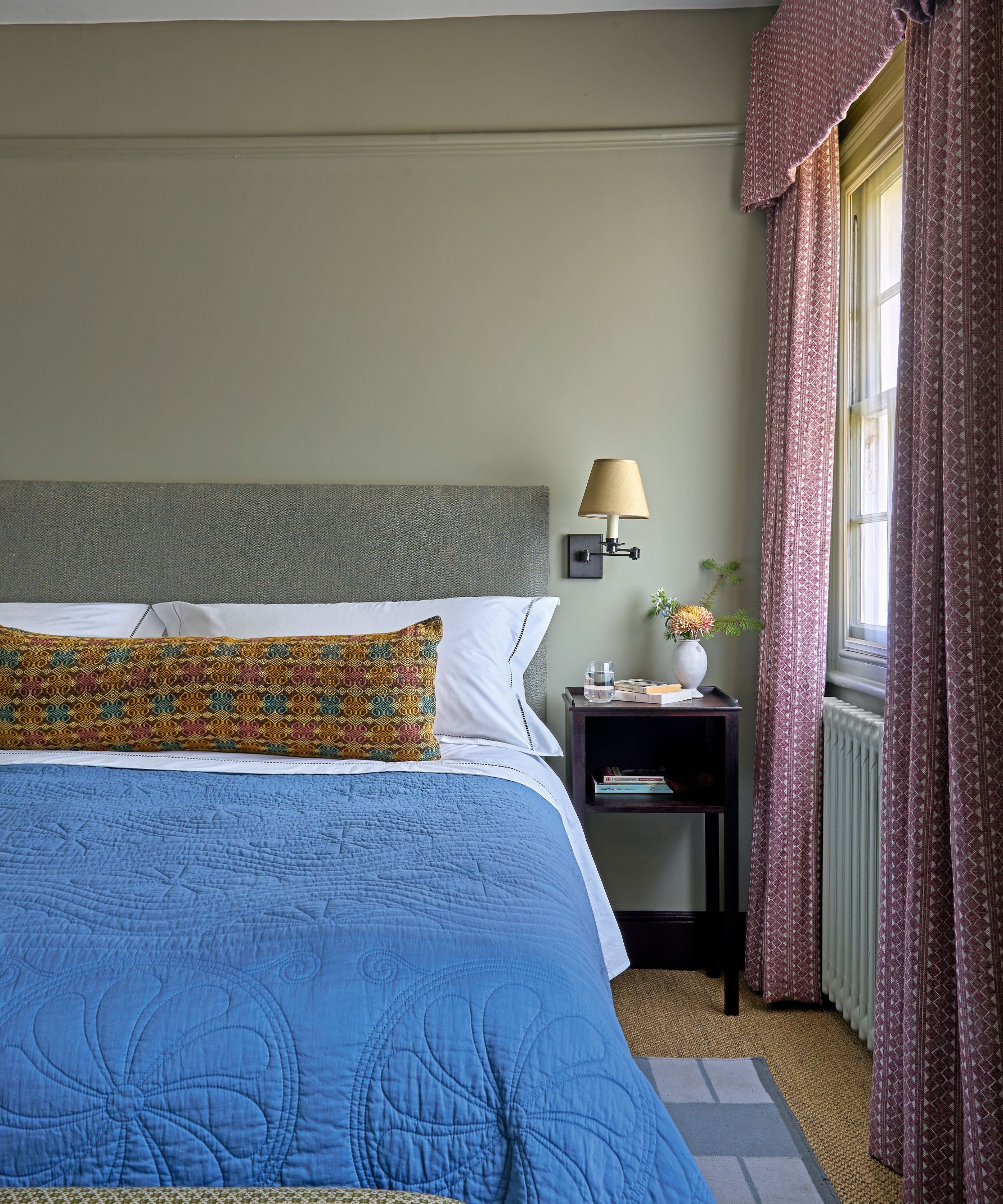 Bedroom with sage green walls, grey fabric headboard, blue quilt and long colourful bolster, rattan carpet and pink pelmet curtains around the window