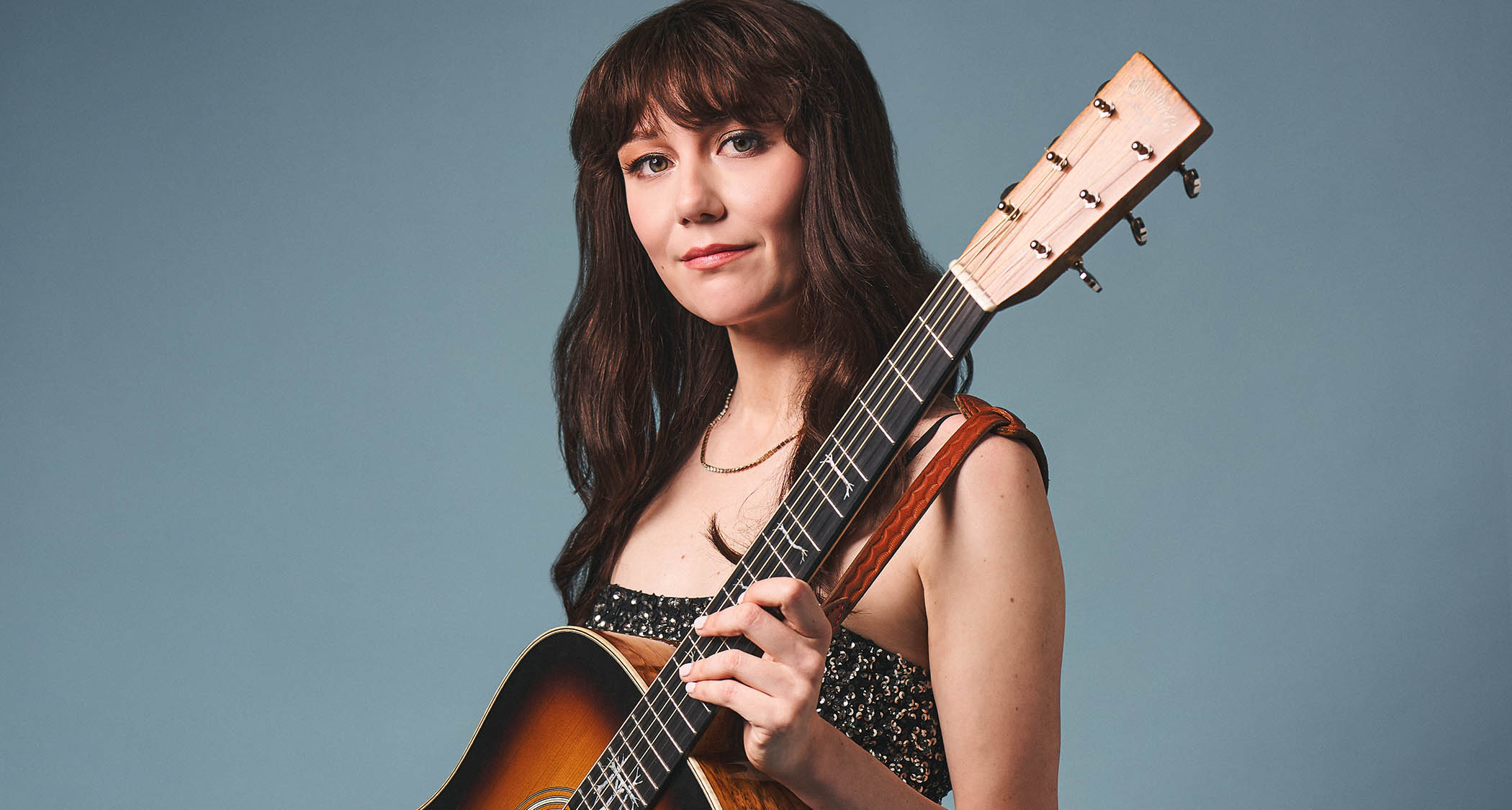 Molly Tuttle photographed with her custom Martin acoustic guitar against a blue-gray background.