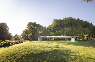 View of MPavilion 2019 by Glenn Murcutt, showcasing its exterior from a distance surrounded by grass and high trees on a sunny day