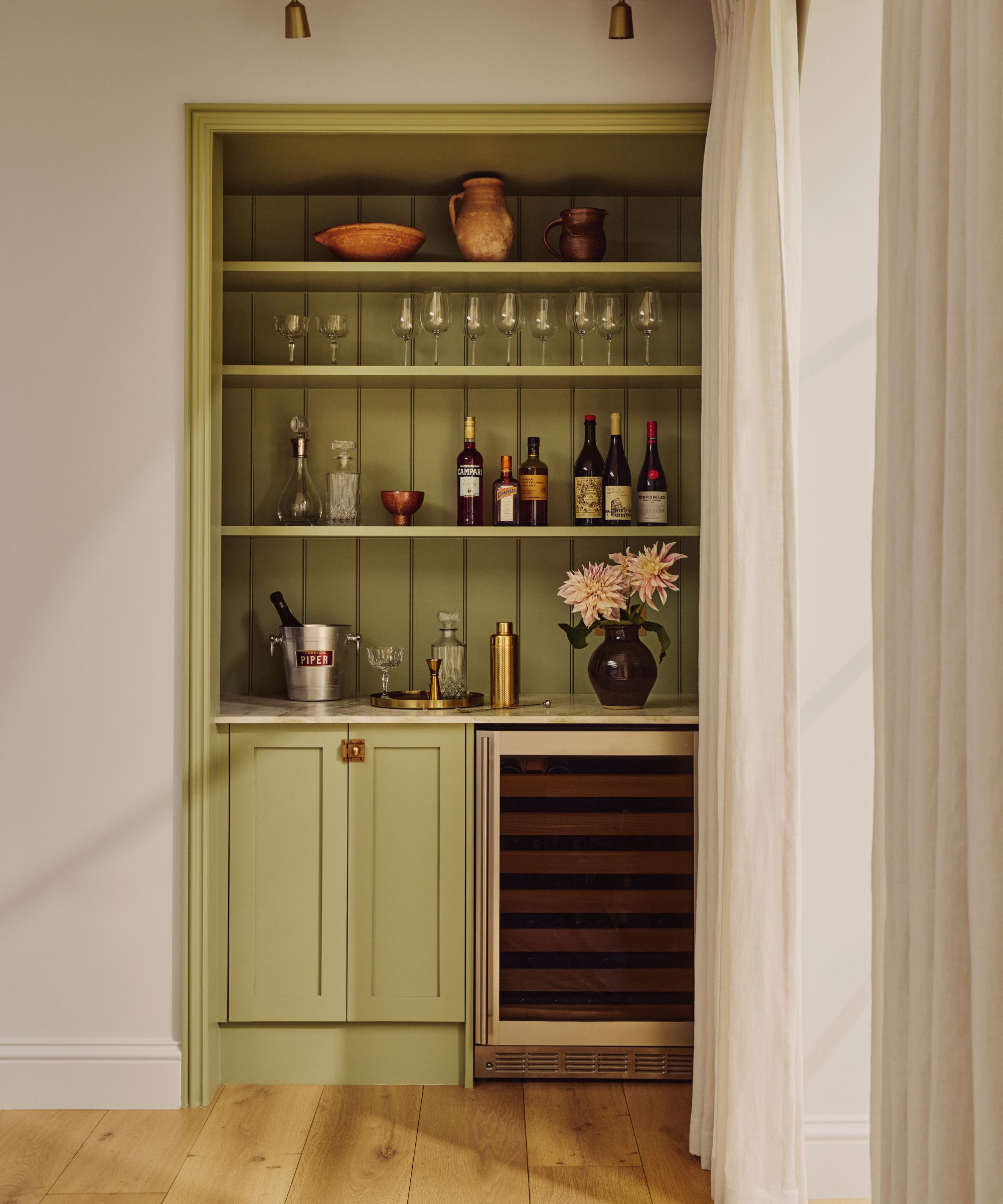 Home bar built into a nook, painted green, with shelves holding glassware and alcohol bottles, a cocktail shaker and ice bucket, and a vase of flowers, with a mini fridge and cupboard below.