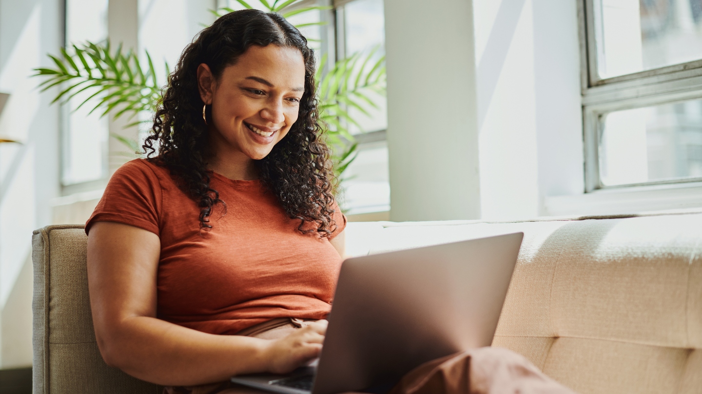 Woman sitting on couch with laptop on lap smiling at her computer