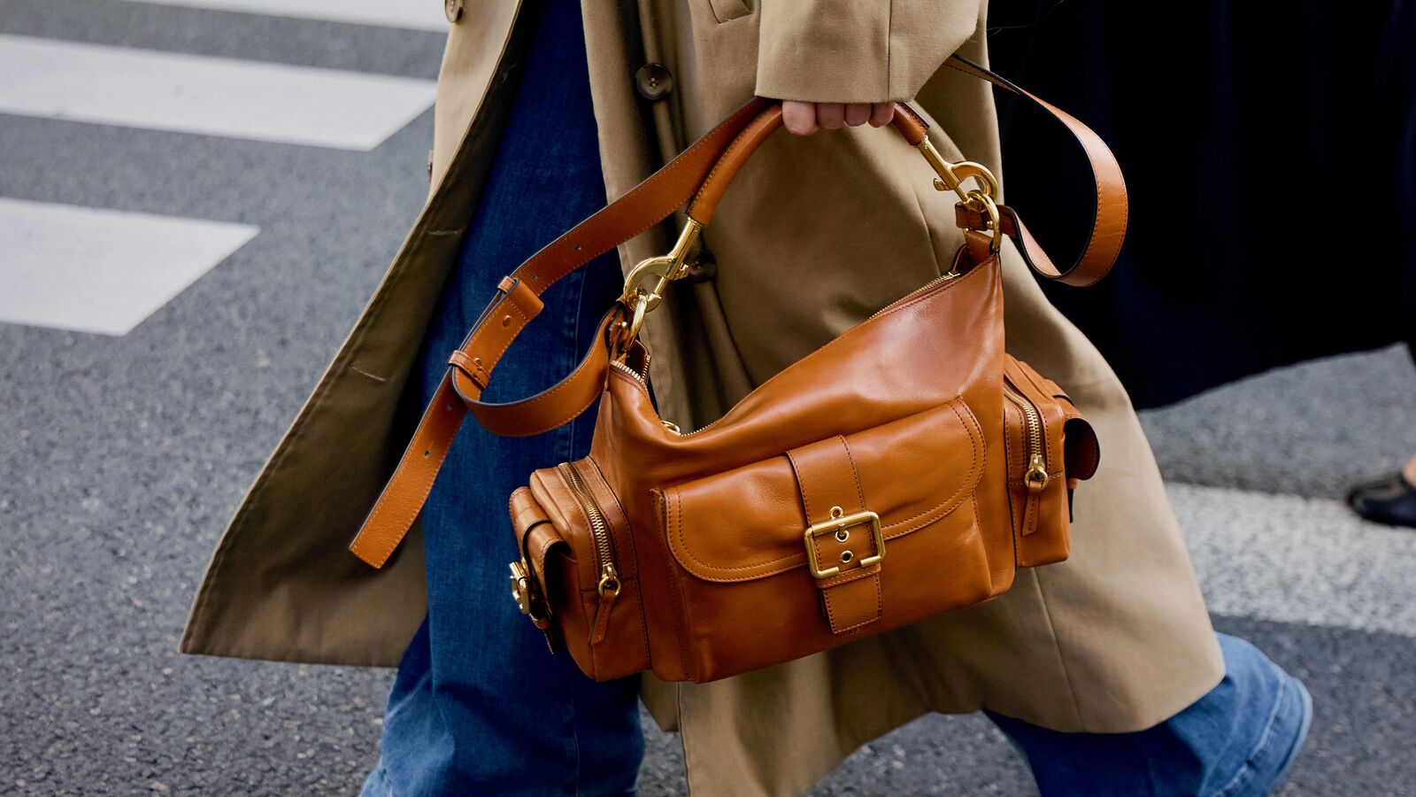 Person in wide-leg jeans, a trench coat, and boots walking in the street holding a leather handbag