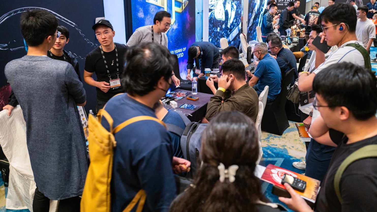 A crowded trade show or convention floor filled with people gathered around tables to test various high-end audio equipment and headphones.
