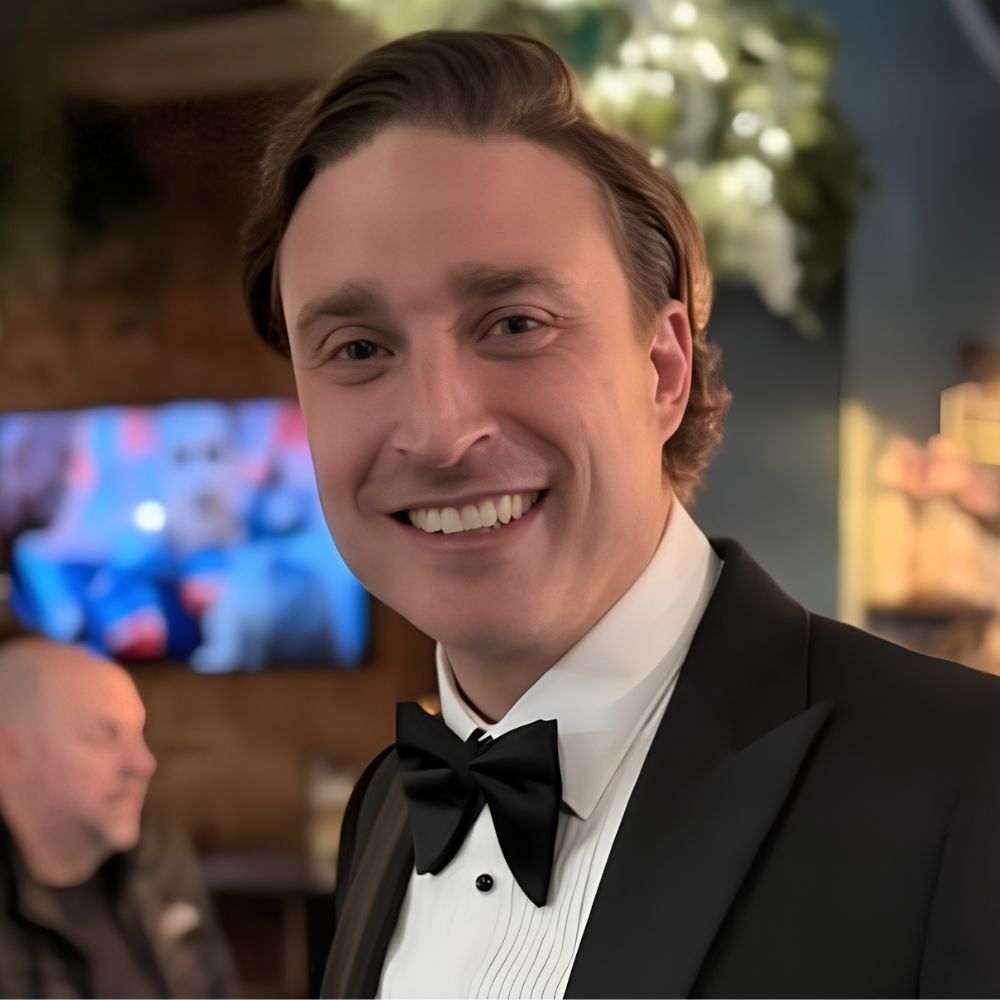A smiling man with brown hair wearing a black tuxedo and bow tie, photographed at a formal event with floral decorations in the background.