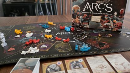 Arcs board game box on top of the board alongside tokens, dice, and cards laid out on a wooden table