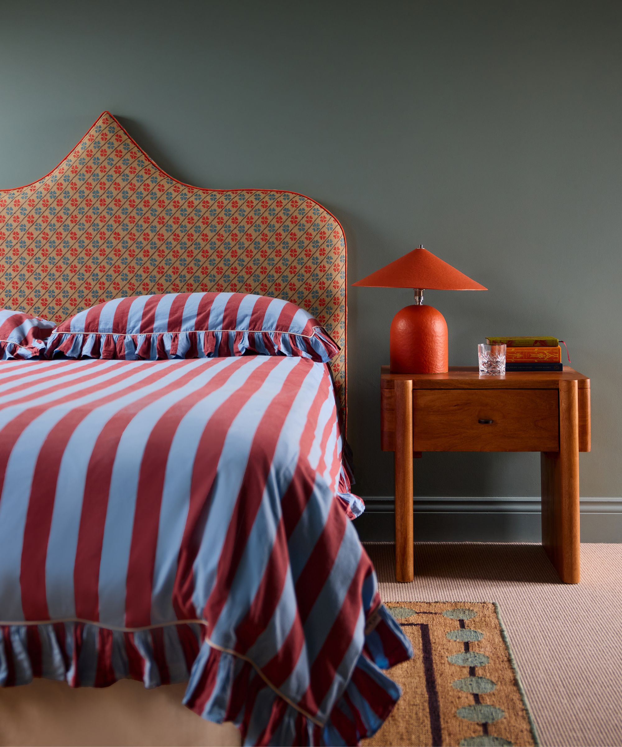 Bed made with red-and-blue striped ruffled bedding and a patterned headboard, paired with a wooden nightstand holding an orange cone-shaped lamp against a dark teal wall.