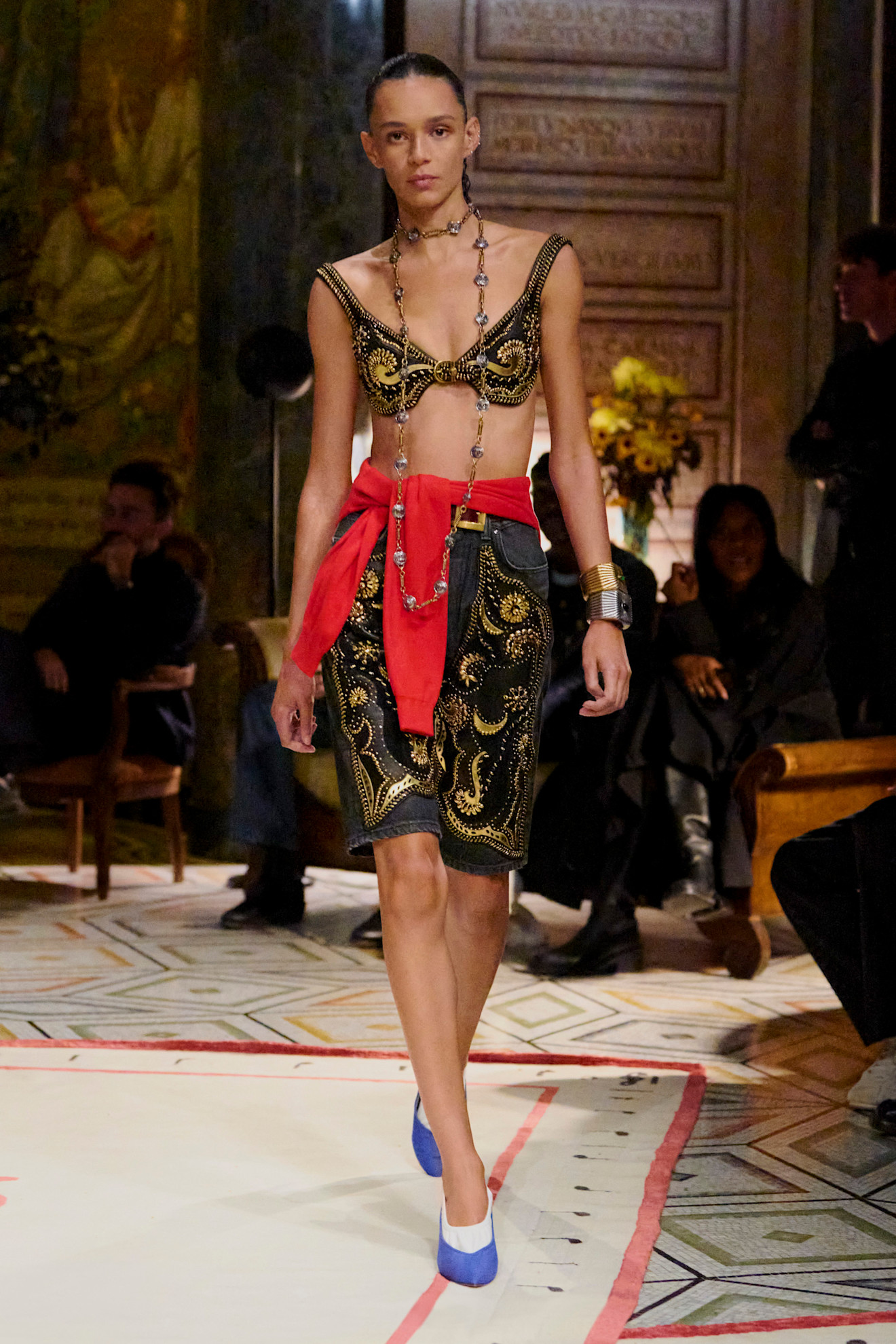 Layering staples for spring 2026 are shown in a photo from Versace's runway show, with a model walking while wearing an embroidered black and gold bra top styled with a matching denim skirt, a red sweater tied around the waist, a long silver and gold chain, gold and silver bangles, white socks, and purple pointed pumps 