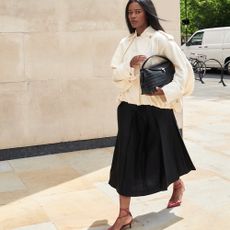 A woman walking in red heels, an ankle-length black skirt, and a beige jacket while carrying a black handbag