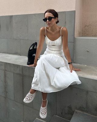 fashion influencer Debora Rosa sits on outside on a concrete wall wearing black sunglasses, a ruched shirred white dress, and crochet flats