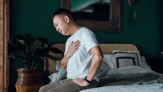 A man sitting on the edge of his bed holding his chest as he experiences heartburn from GERD