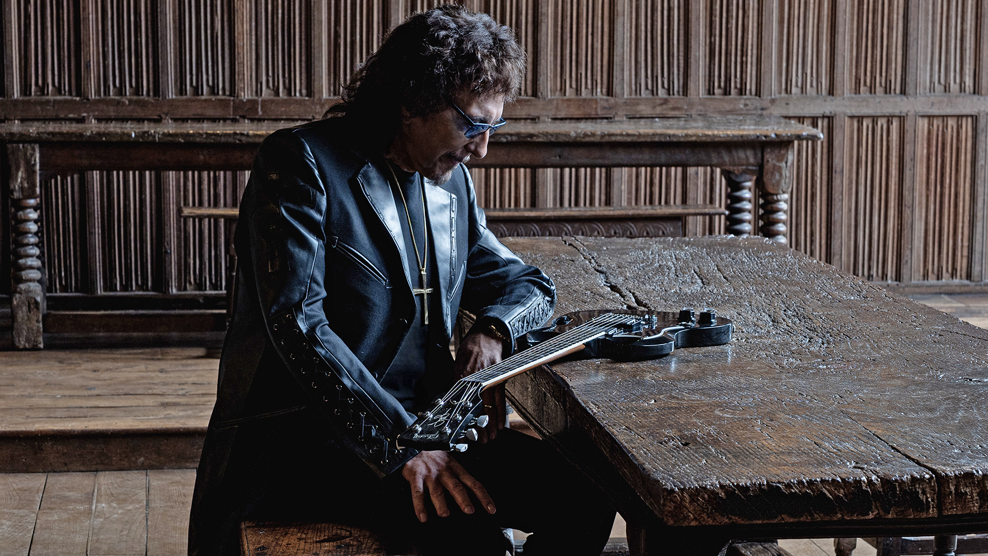 A photo of Tony Iommi staring at a left-handed Gibson SG resting on a table in a room with brown paneling. Issued in conjunction with the announcement of his 2026 solo album, From the Dark.