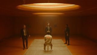 Image from the Marvel T.V. show Loki, season 2 episode 2. Sitting on a chair is a man in a white jumpsuit and he is surrounded by three men in suits standing over him and interrogating him.
