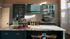 A kitchen with muted blue cabinets, large island with gold faucet, wooden chairs, fruit on wooden board and white veined gray marble countertops