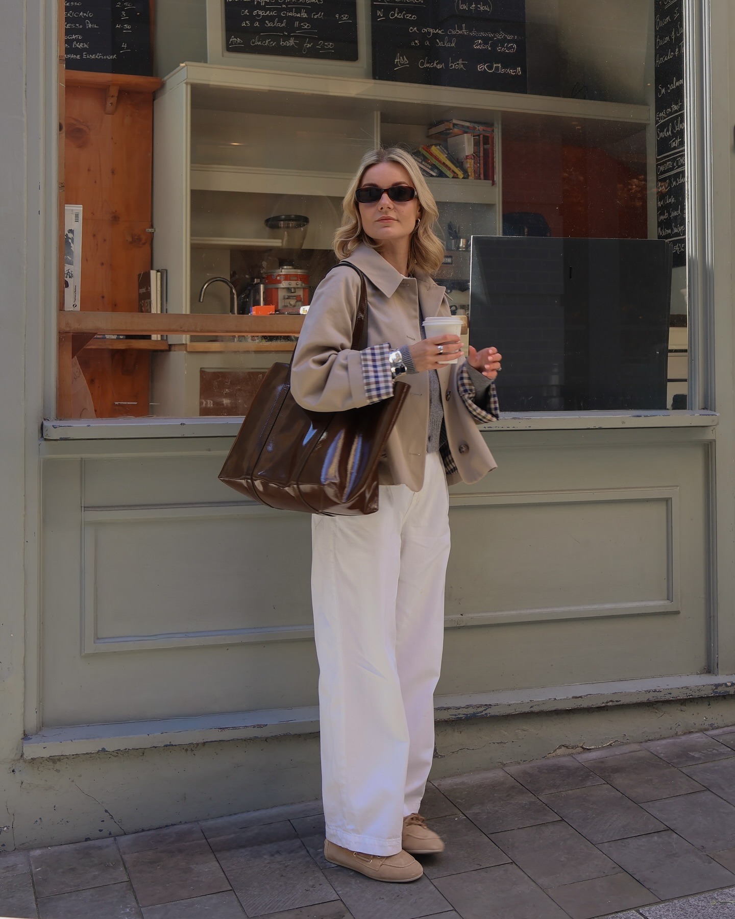 Fashion person Nina Caine wears the best autmn jacket in a cropped trench. She pairs her trench with white jeans a vinyl bag, suede loafers and a grey cardigan. She is stood in front of a sage green coffee shop looking past the camera in a recent image from her Instagram.