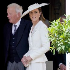 Catherine, Princess of Wales emerges from Windsor Castle wearing a white lace-trim skirt with a tonal jacket neatly fastened around her waist. She wear a wide-brim white hat, dark beige heels and carries a small satin clutch in her hands.