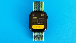 Detail shot of an Apple Watch SE on a bright blue background showing the steps to set up and send a Check In notification in watchOS 11 