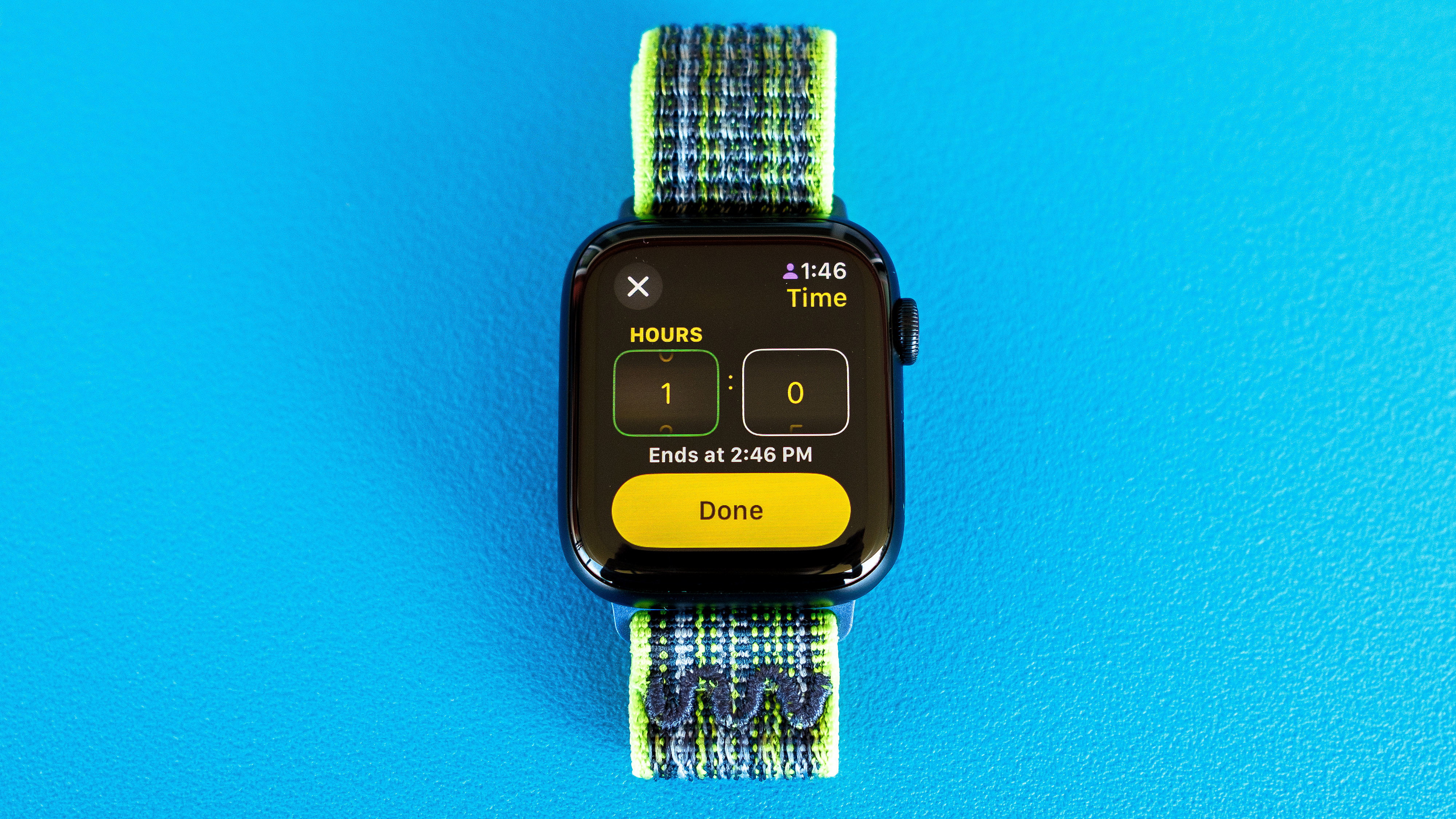 Foto detallada de un Apple Watch SE sobre un fondo azul brillante que muestra los pasos para configurar y enviar una notificación de Check In en watchOS 11 