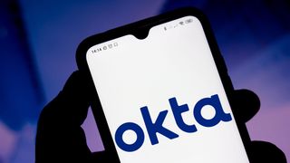 Okta logo appearing on a smartphone held aloft by a silhouetted hand in front of a blue and purple background