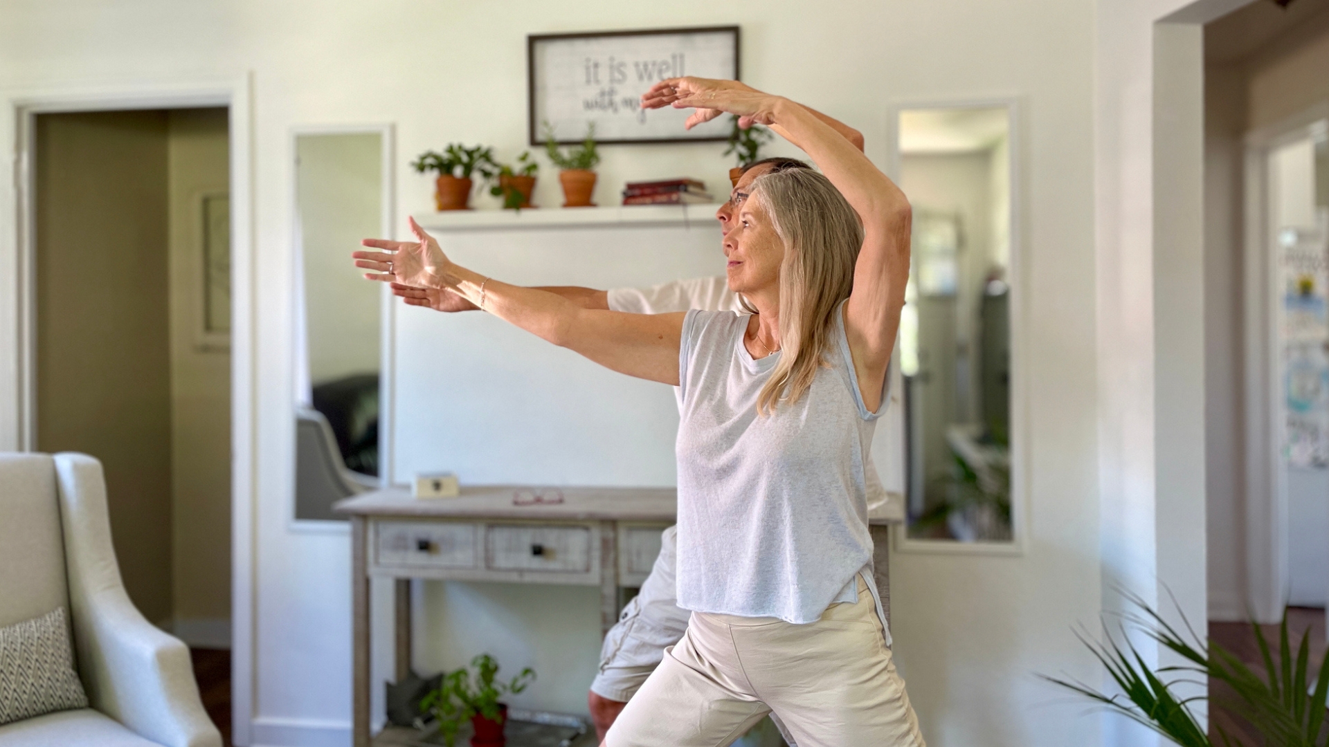 woman standing in front of a man both performing tai chi moves in a living room setting looking sideways away from the camera