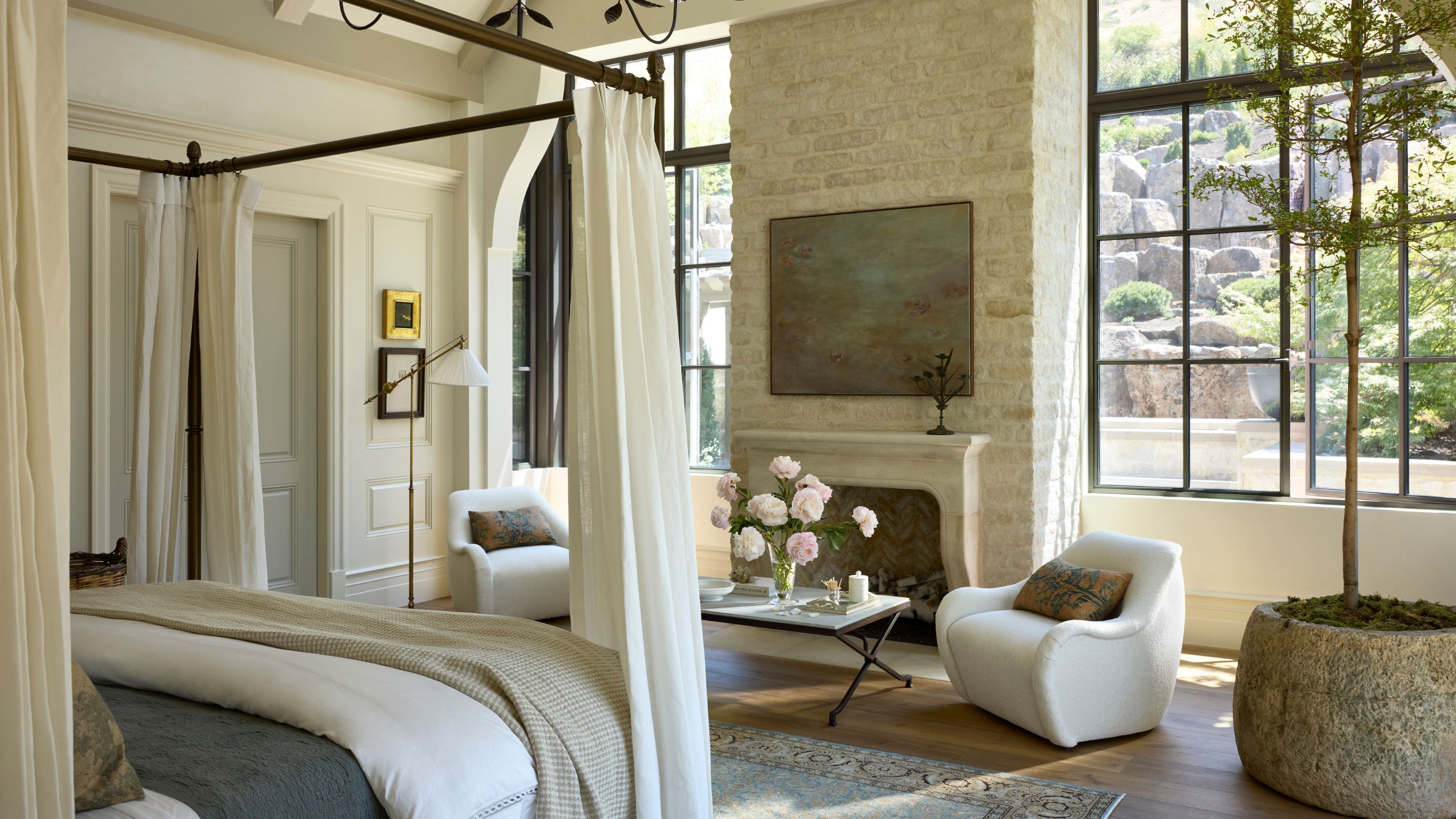 a large bright primary bedroom with vaulted ceiling, large windows, stone fireplace, four poster bed and sitting area