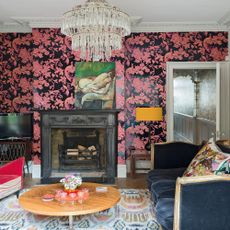 Living room, bold, dramatic pink and black wallpaper with birds of paradise motif pattern, round circular vintage coffee table, rug, blue velvet French sofa, wrought fireplace, crystal chandelier 