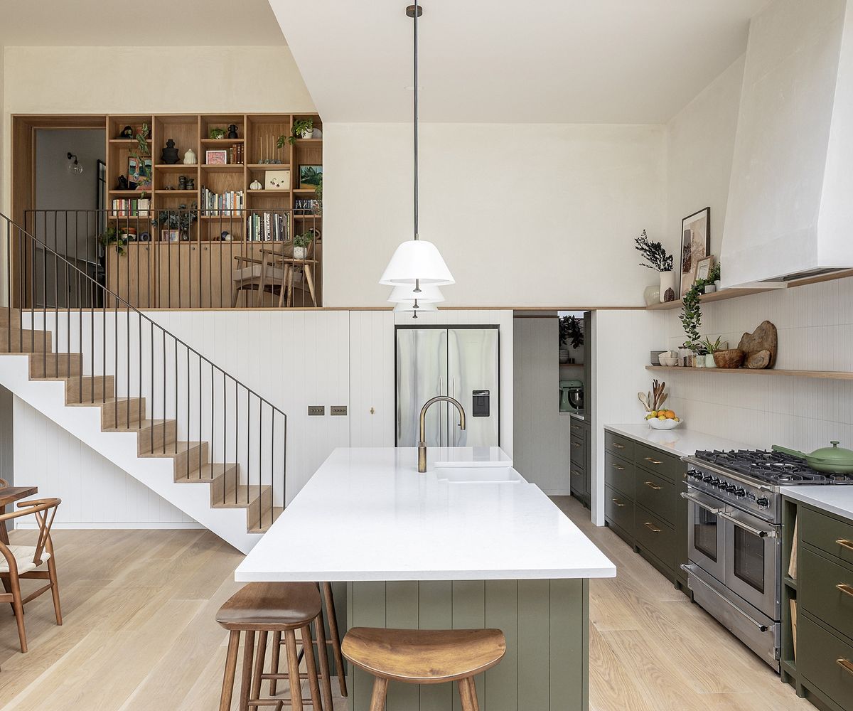 kitchen with dark khaki cabinets and light surfaces and tiles. There are three pendant lights and a light oak floor. There is a staircase on the far wall leading up to the next floor