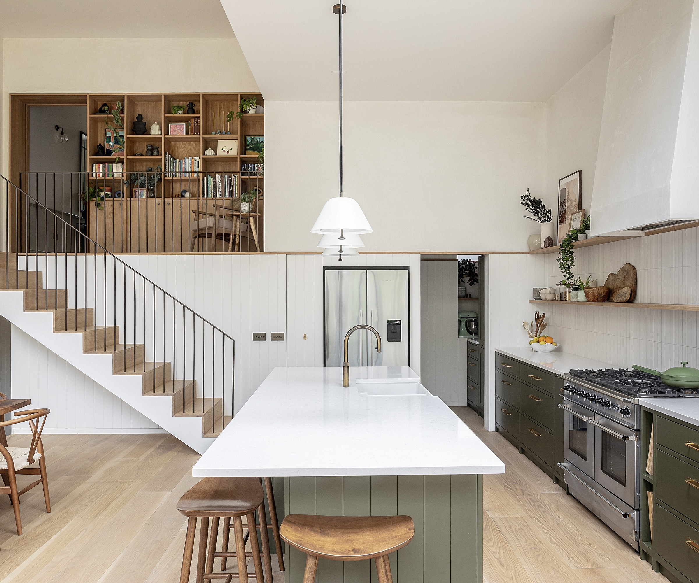 kitchen with dark khaki cabinets and light surfaces and tiles. There are three pendant lights and a light oak floor. There is a staircase on the far wall leading up to the next floor