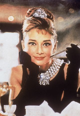 Audrey Hepburn in a black, shoulderless dress, matching gloves, and a tiara, smiles with a cigarette holder in her hand, in her role as Holly Golightly the film 'Breakfast at Tiffany's,' directed by Blake Edwards, New York, New York, 1961