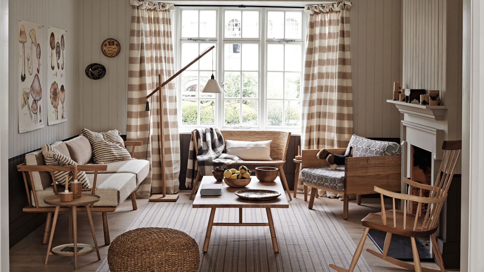 A neutral living room with wall panelling, a striped rug, wooden table, wooden chair, brown and white gingham curtains over a large window, and a white mantelpiece.