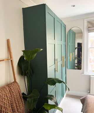 Bedroom with dark green painted wardrobe, light wooden ladder with throw, arch mirror on wall beside mirror, cream carpet, plant beside wardrobe